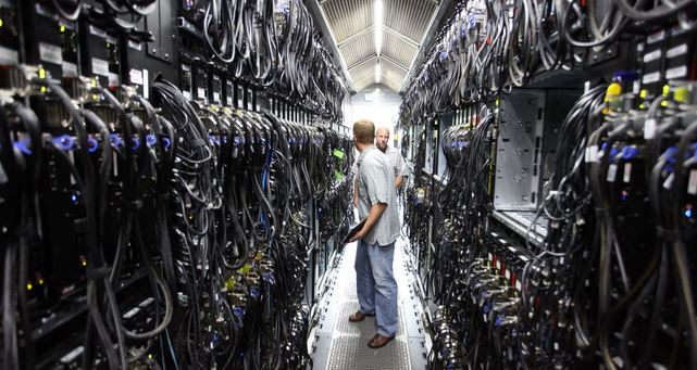 Picture of two guys standing in an aisle between rows and rows of complex computer servers in a massive data center