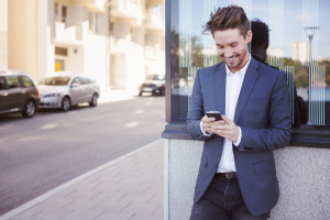 Young businessman using mobile phone against wall by sidewalk