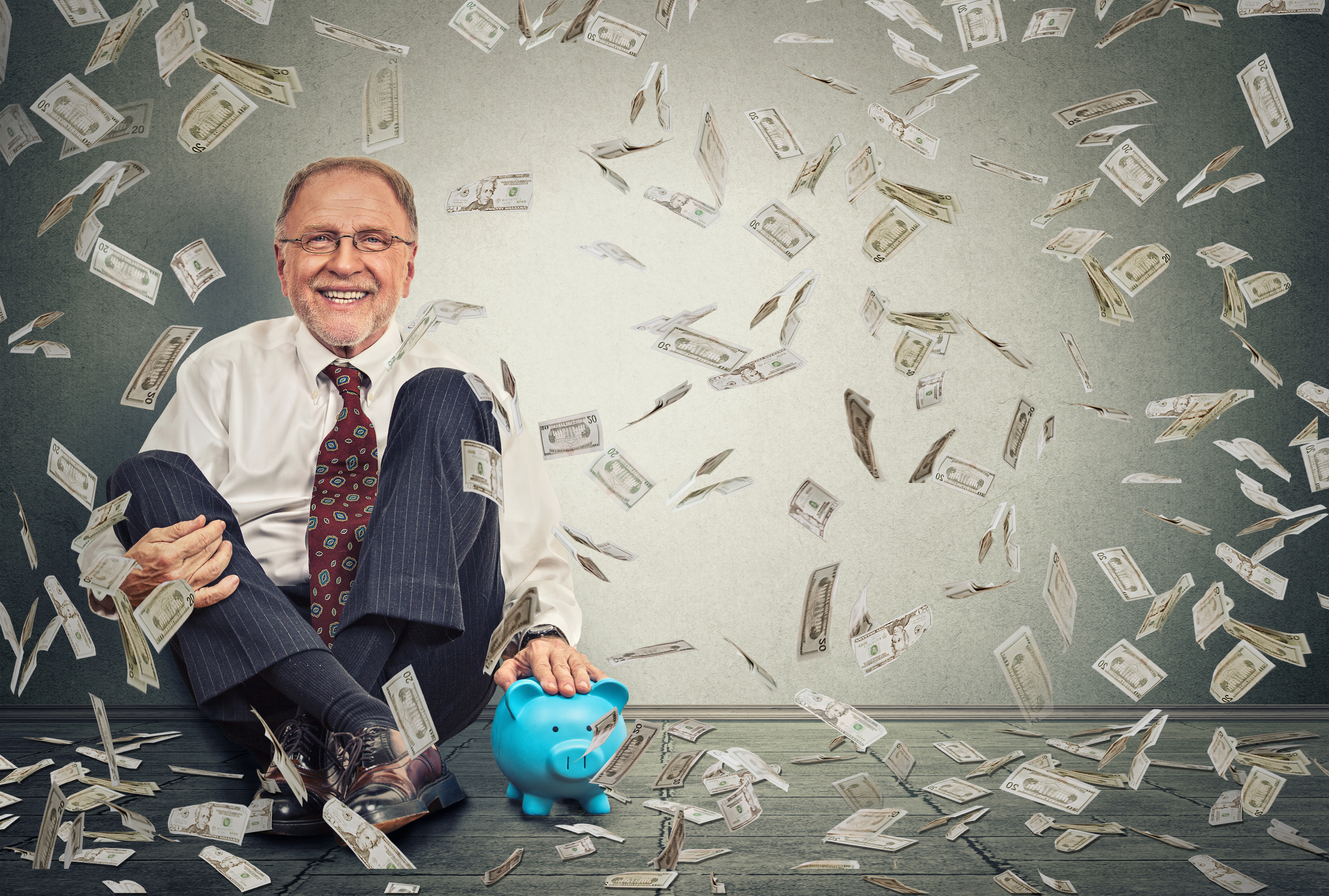 Excited happy senior man sitting on a floor with piggy bank under a money rain isolated on gray wall background. Positive emotions financial success luck good economy concept