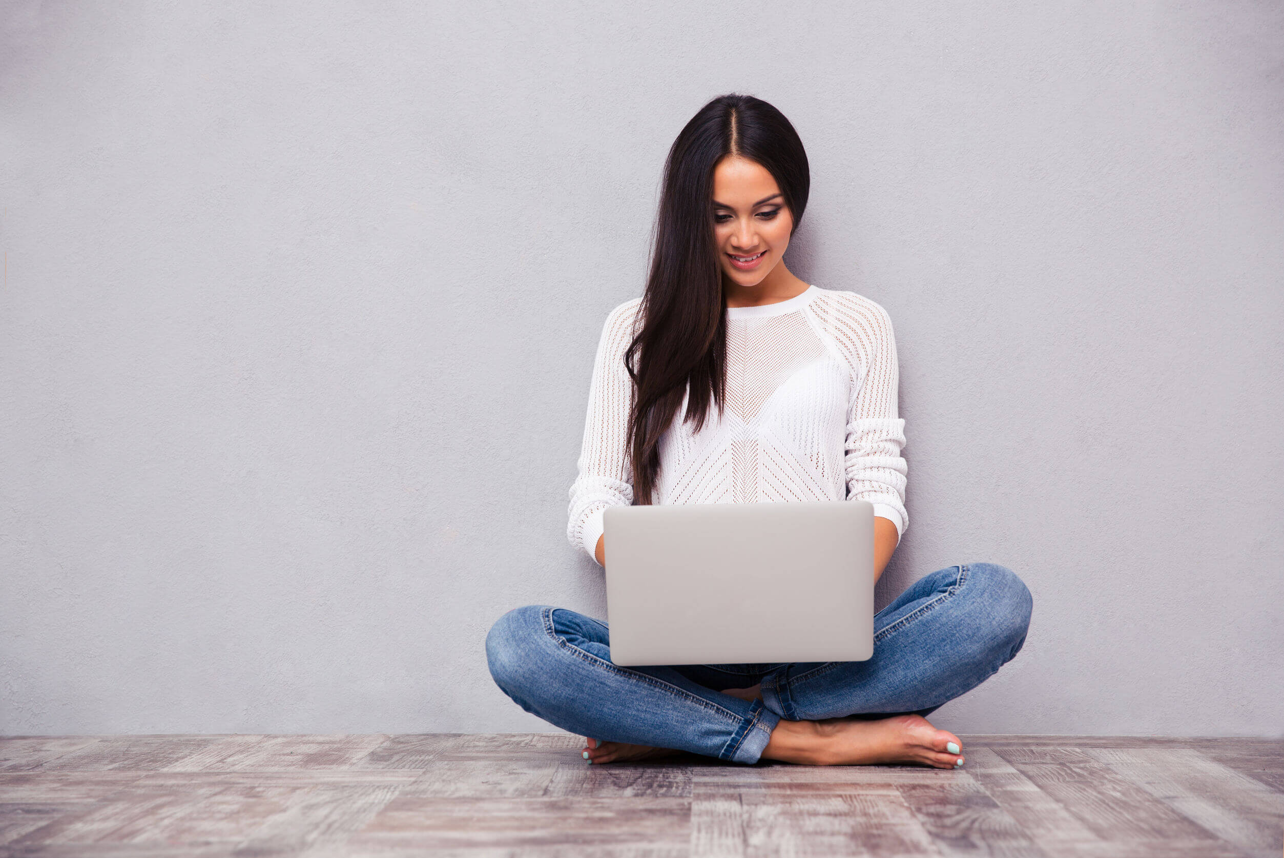 portrait of a happy casual woman sitting on the floor with laptop on gray background