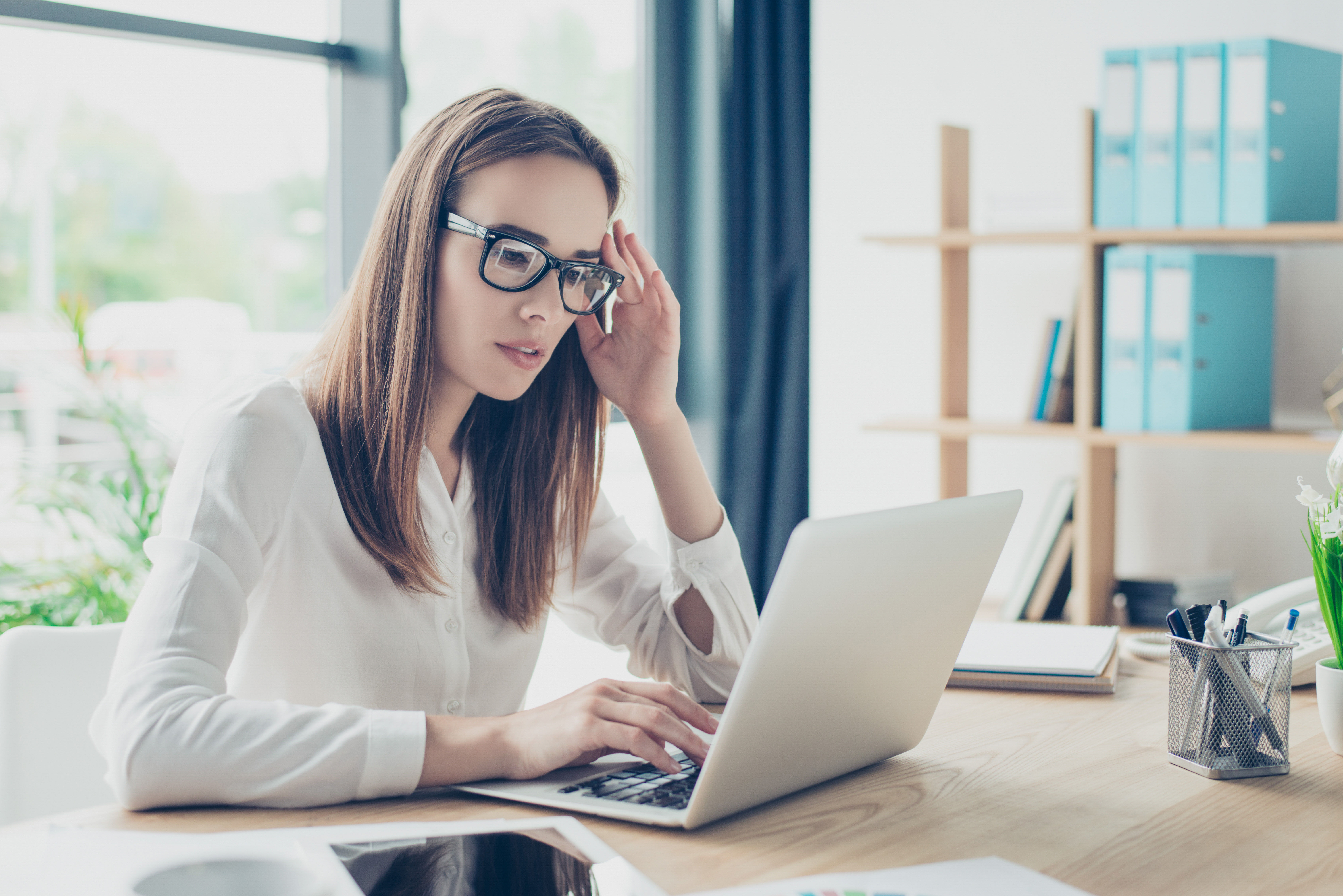 Close up portrait of successful business woman economist in formal wear, sitting at her work place in a light modern design office, sitting in front of laptop, posting social media and looking pensive.