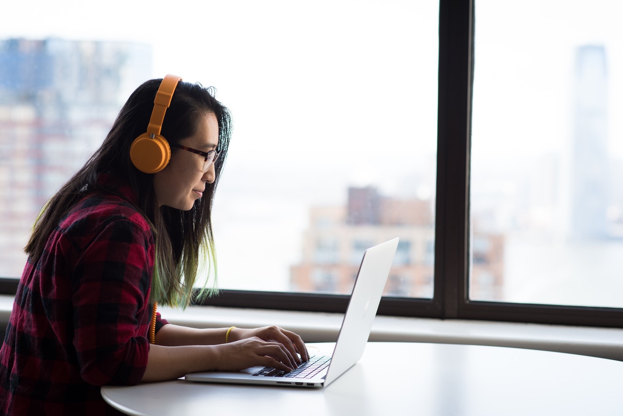 A woman sits at an empty table on her laptop. She is wearing headphones. The background is a window overlooking a city.