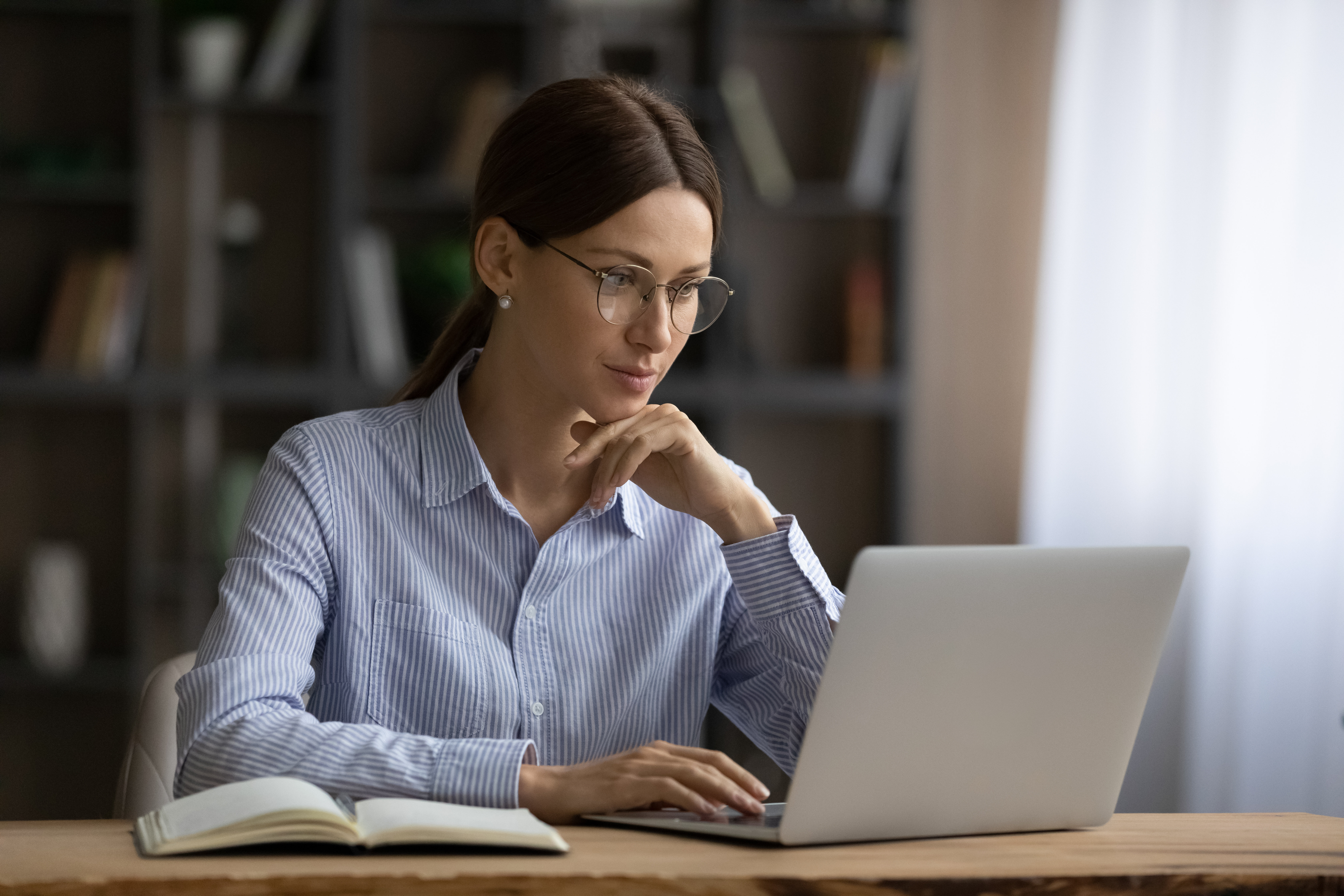 Online content creation for business. Focused young lady in glasses sit at home office engaged in computer job read information on laptop screen. Thoughtful millennial woman chat in corporate network type email text on pc
