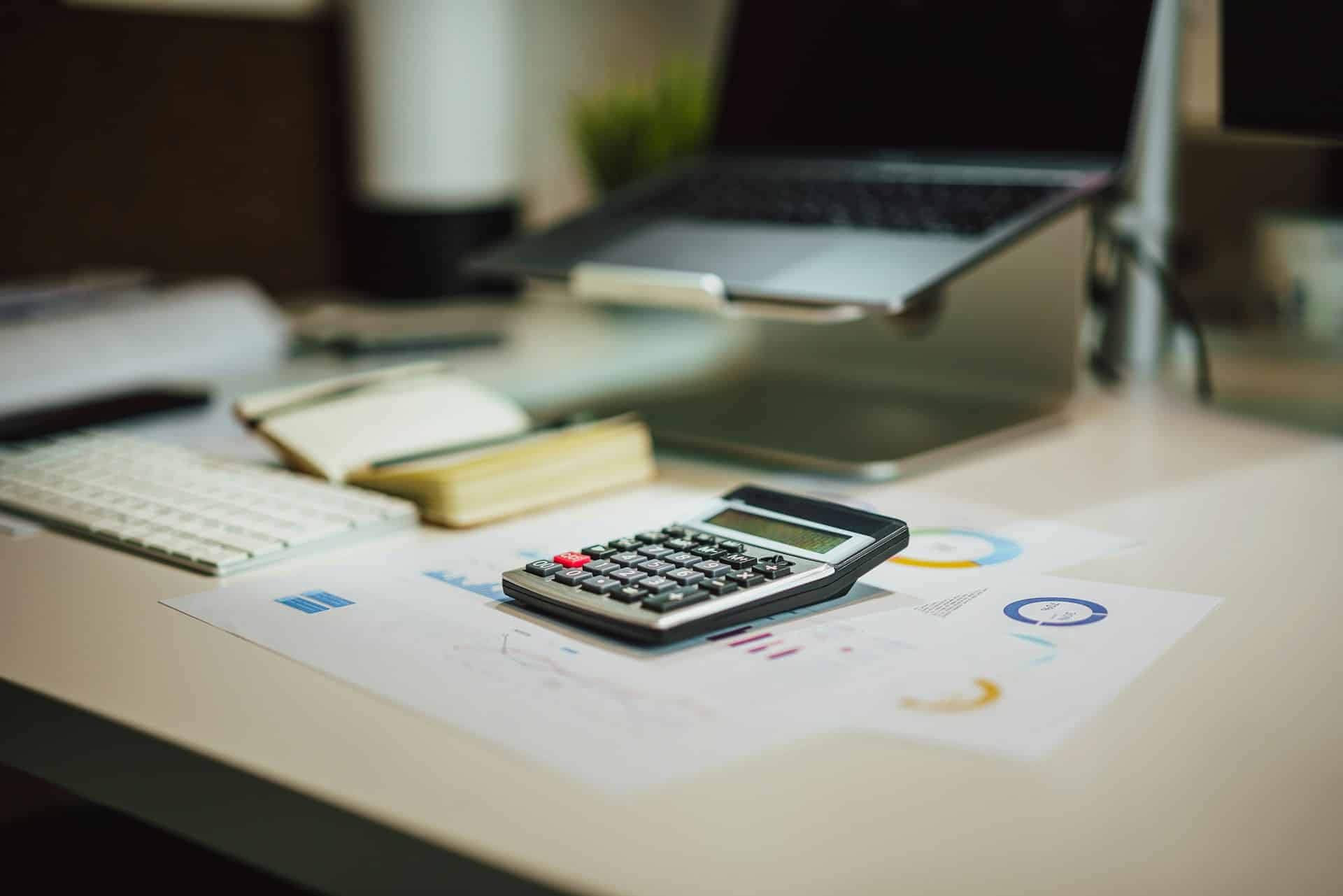 A calculator sits on top of printed financial charts on a desk beside a laptop, keyboard, and notepad.
